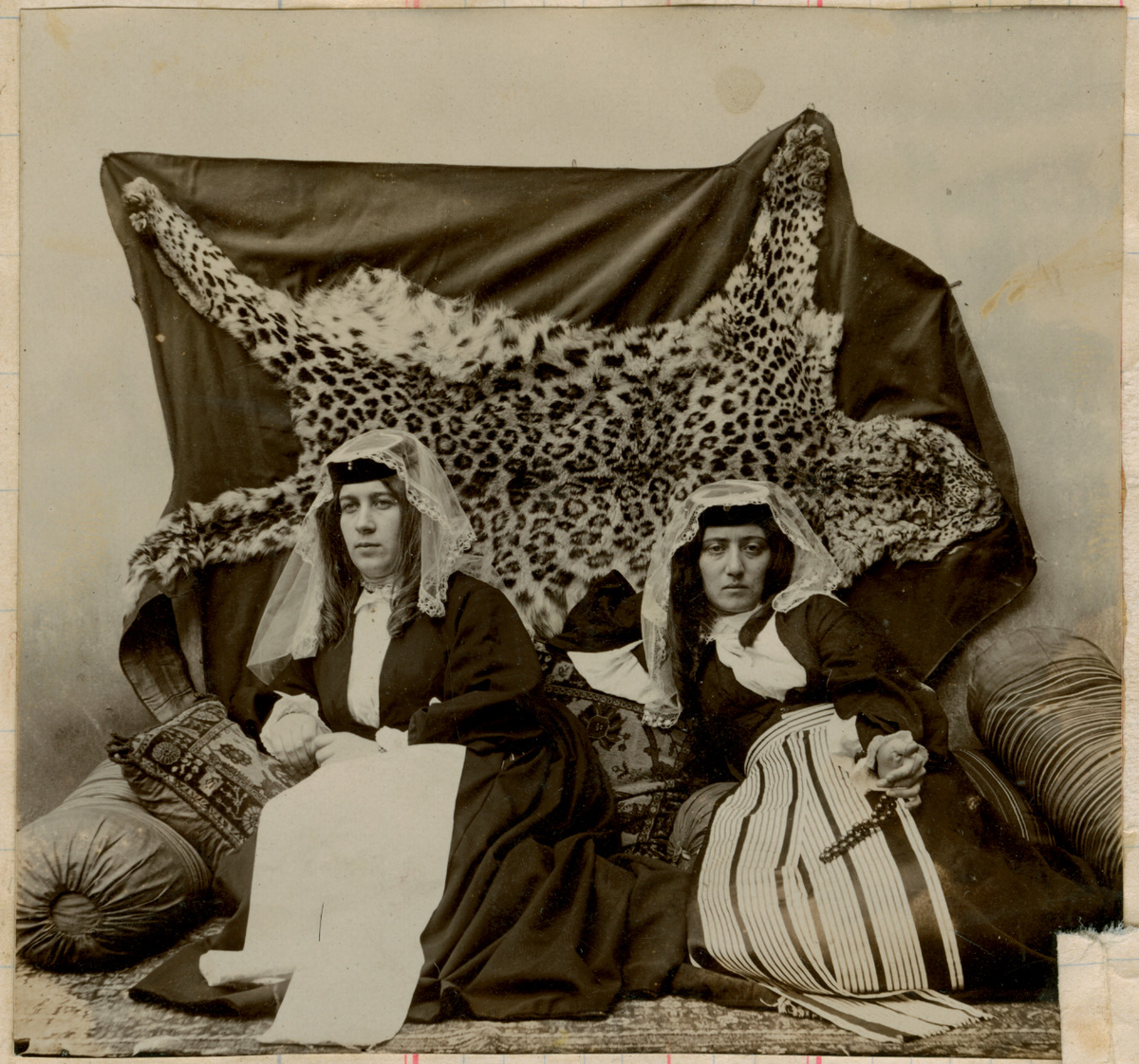 Studiofotografi av to kvinner liggende på et teppe med puter. Begge kvinnene bærer slør. Kvinnen til høyre holder et perlesmykke. På veggen bak henger et leopardskinn.