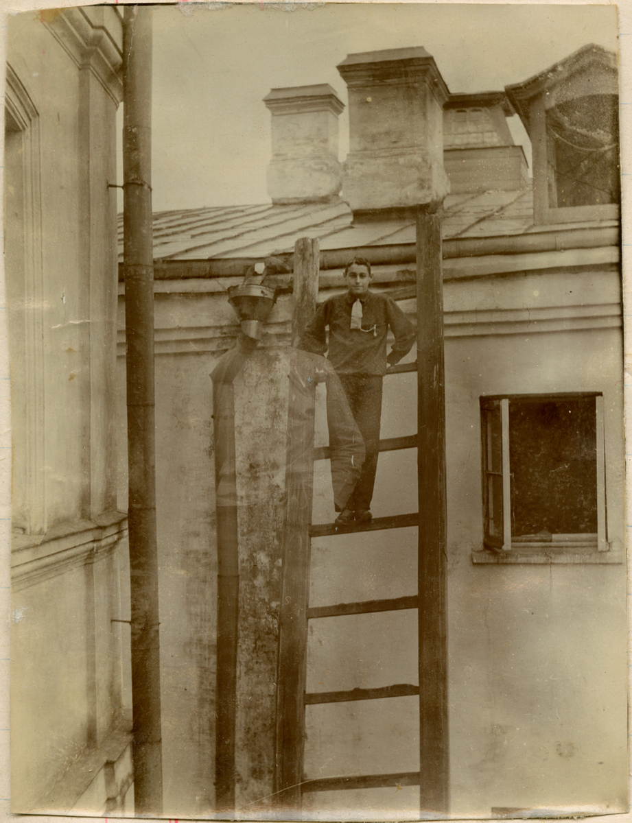Motivet viser en ung mann poserende øverst i en trestige. På taket ses tre skorsteinspiper, nedløpsrør og et åpent vindu til høyre på husveggen.