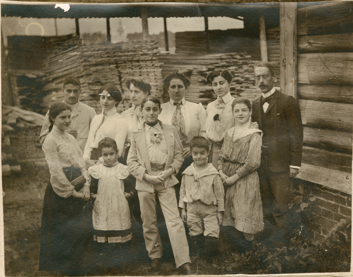 Motivet viser familien Rostin samlet for fotografering foran en tømmerbygning. Trolig teglsteinfabrikken i Borovitsji. Mennene på bildet er ukjente.