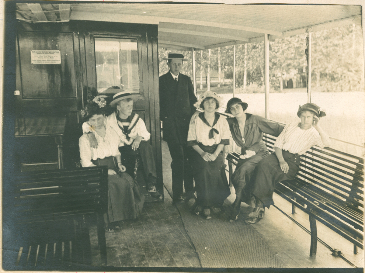 Et utendørs gruppebilde av to menn og fire kvinner på noe som kan være en elvebåt. De har på seg sommerlige klær og hatter. Noen med maritimt tema.