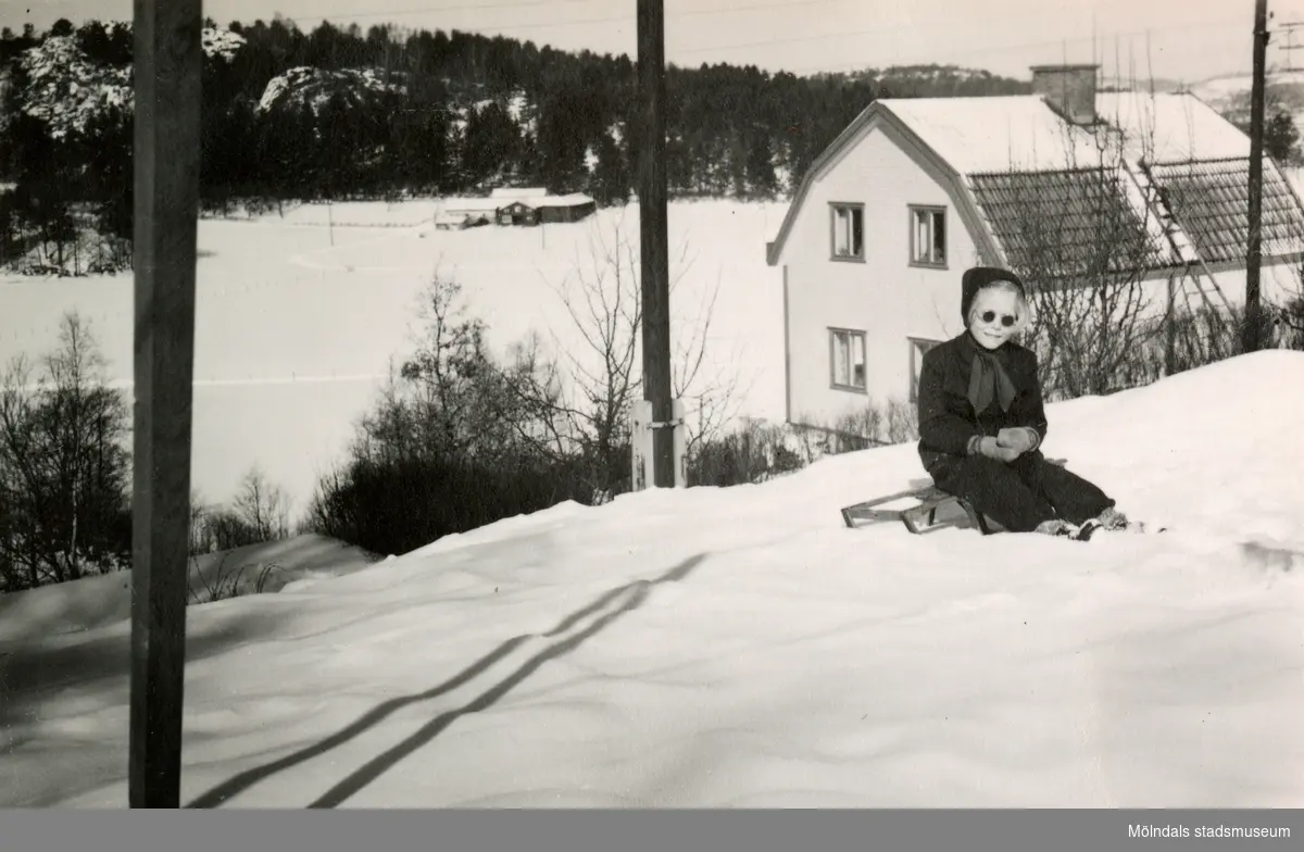 Eva Pettersson (född 1944, gift Kempe) sitter på en kälke framför bostaden på Gamlehagsvägen (ej i bild) år 1951. I bakgrunden ses Alvered.