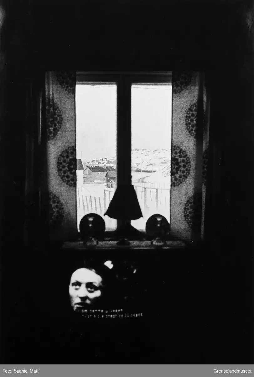 Parti fra Bugøynes sett ut gjennom et vindu. En tv står på i forgrunnen. På et konsollbord foran vinduet står en lampe og to lysestaker.