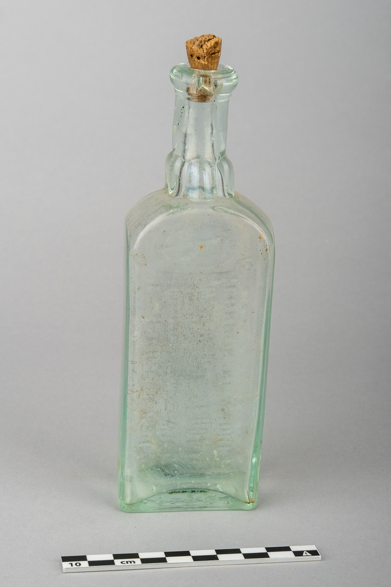 Flaske av blankt glass med svakt grønnaktig skjær. Rektangulær form med skrift på den ene bredsiden.