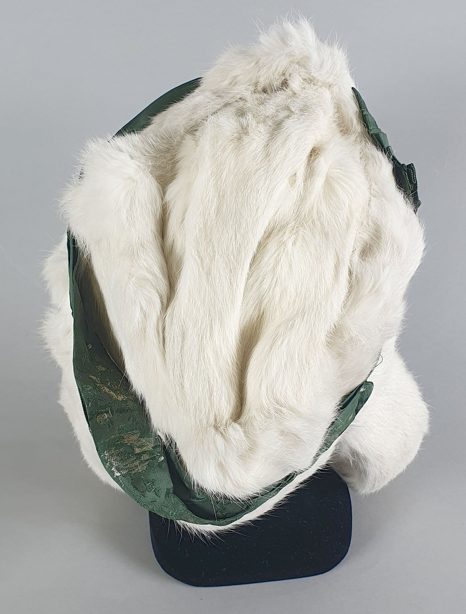 Hvit pelslue av fjellrev eller kanin, med pelsbånd på hver side. Grønt brokadbånd rundt pullen med en rosett på siden, Fôret med gas og strie.
