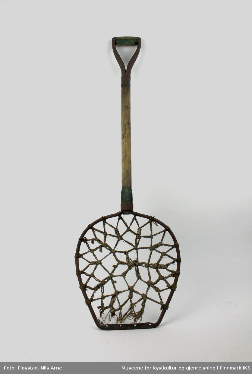En sildespade med en oval ramme av jern med et nett av hampetau festet til rammen. Skaftet er av tre med et håndtak av både jern og treverk.