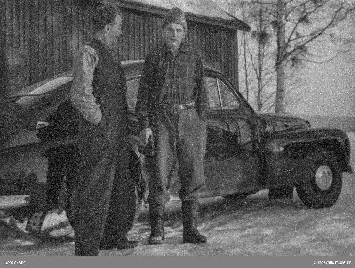Två män som står invid en blank Volvo PV, bakom bilen en husvägg, lantlig omgivning och snö på marken. Ur Anders Nylanders samling.