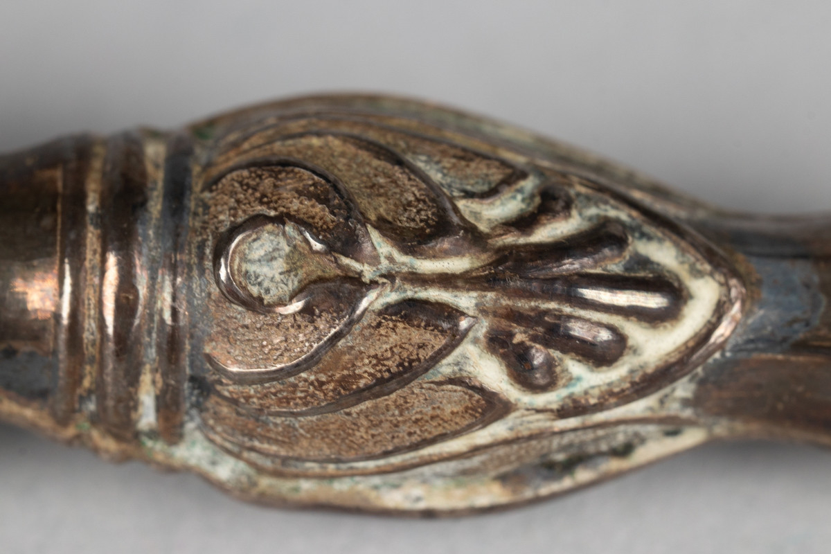 Etui av sølv, avlangt, sylinderformet med blad ornamentikk i  endene. Lokk hengslet fast med lenker.
