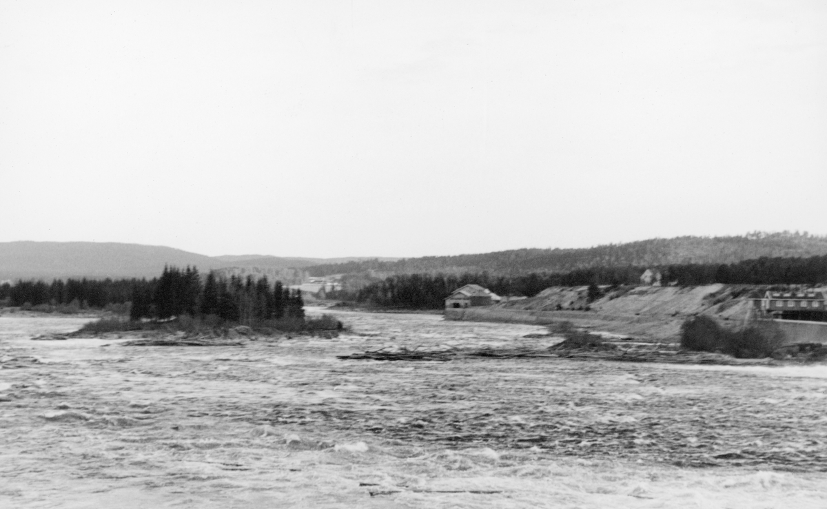 Utsikt over Glommas løp nedenfor kraftverksdammen ved Skjefstadfossen i Heradsbygda, Elverum i Hedmark. Bildet ble tatt i juni 1941. på et tidspunkt fa det var flom i elva. Fotografen har stått på elvebrinken på østsida av vassdraget med kameraet vendt sørvestover langs elveløpet, som her er ganske vidt. Vi ser at en del tømmer hadde pakket seg sammen i en haug på ei steinør i den midtre, grunneste delen av elveløpet, mellom dammen og Tjuvholmen. Helt til høyre i bildet skimter vi den vestre delen av kraftverksdammen. Sentralt i bildet ser vi kraftstasjonen ved vestre elvebredd, cirka 400 meter nedenfor dammen.
