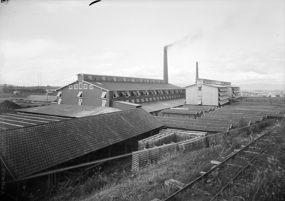 Strinda Teglverk. Fabrikkbygninger i bakgrunnen og rader med teglstein fremfor bygningene. Foran fabrikken vises et jernbanespor.