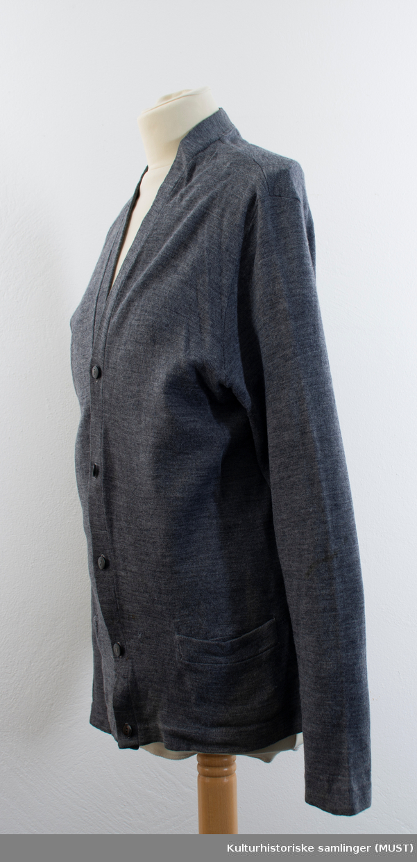 Strikkejakke i mørk grå tynn ull med V-hals og knappestolpe med svarte gråknapper. Nederst framme på jakken er det en lomme på hver side av stolene. 