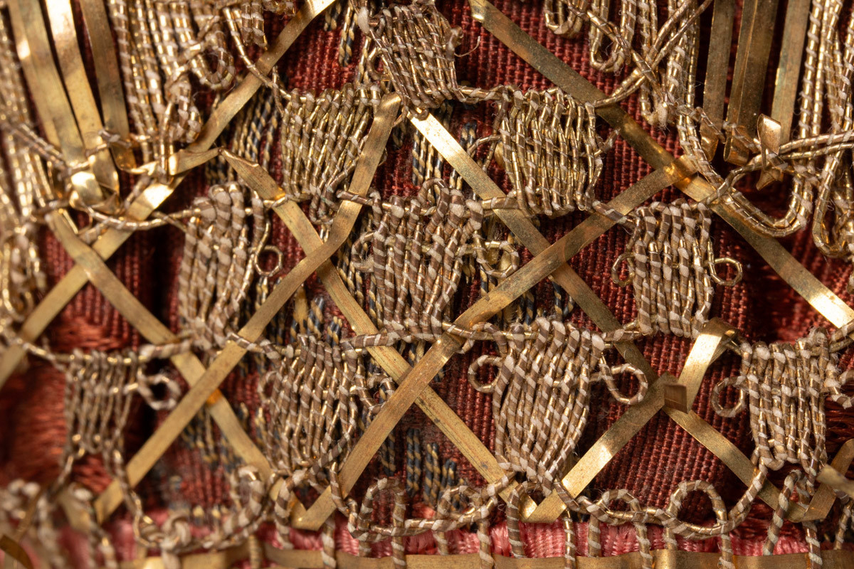 Dåpslue i brun silkebrokade med gullkniplinger og kanting og bånd i rosa silketaft. Luen er satt sammen av tre deler, og har lett mønstret foring i lin.