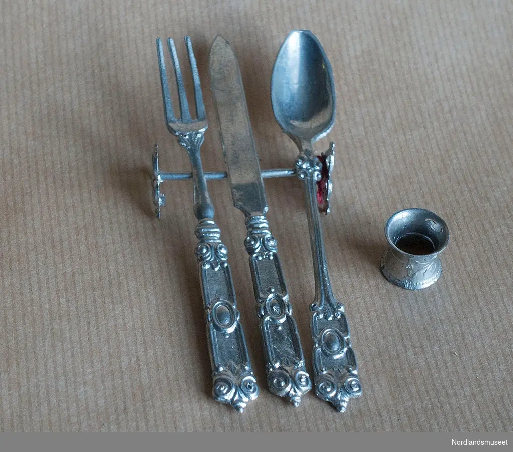 Lekebestikk i fem deler: skje, gaffel, bordkniv, serviettring og bestikkholder, alt utformet som voksenbestikk, antatt fra rundt 1900