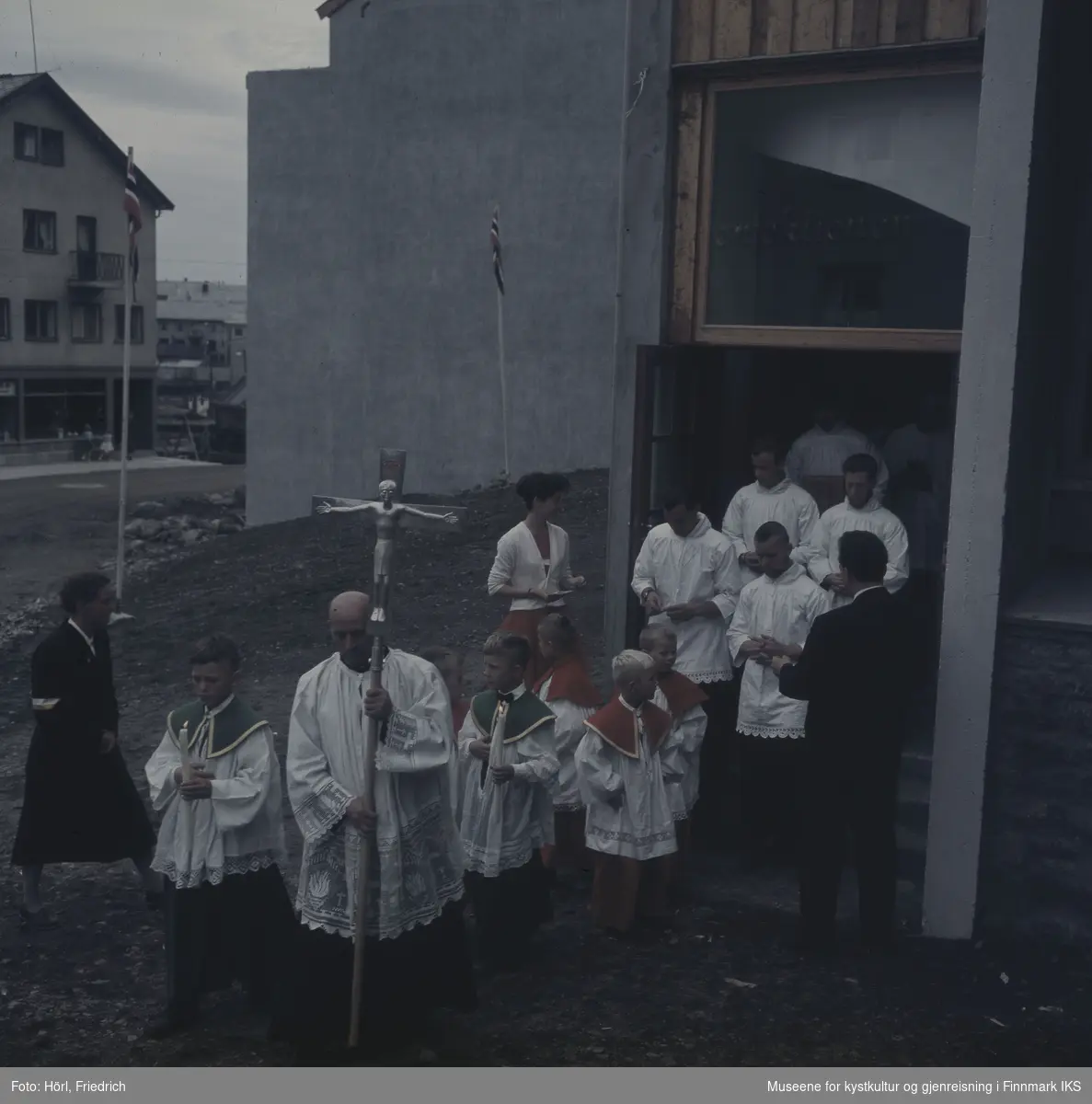 Presten bærer prosesjonskorset og leder an toget ut av Den katolske kirke St. Mikael i Hammerfest i 1958. Ministrantene går bak ham og to av dem holder stearinlys.