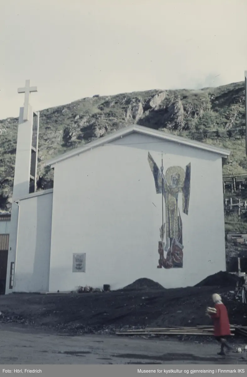 Den nybygde katolske kirke St. Mikael i Hammerfest står ferdig en solskinnsdag i 1958. Kirka er fotografert fra Strandgata og man ser Salenfjellet i bakgrunnen. Kirkens yttervegg er dekorert med en stor mosaikk som viser St. Mikael. Mosaikken er skapt av den tyske billedkunstneren Karl Manninger. En kvinne i rød kåpe med en pakke i armen går forbi.