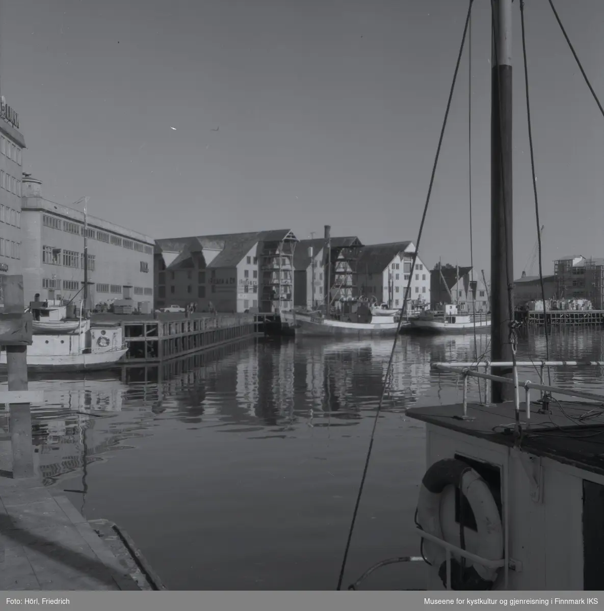 Bildet er tatt i Tromsø havn der fiskebåter ligger til kais. I bakgrunnen er det pakkhus og industribygg. På et av bygningene står det "Rieber & Co."