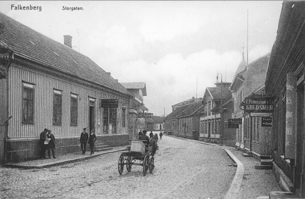 Vykort, "Falkenberg Storgatan." Stadsbebyggelse med bostadshus och butiker. Till höger liger Petterssons Guldsmedsbutik och på gatan kör en hästdragen kärra. Vid fasaden till vänster står ett par män och läser tidningen. Årtal och fotograf är okänt.