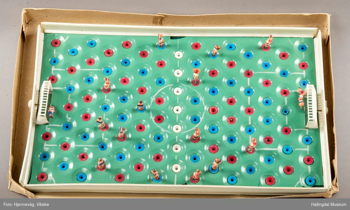 Fotballspill med grønn matte, hvite mål m/målmenn. 6 røde og 8 blå spillere. Det mangler flere spillere. Ligger i orginaleske.