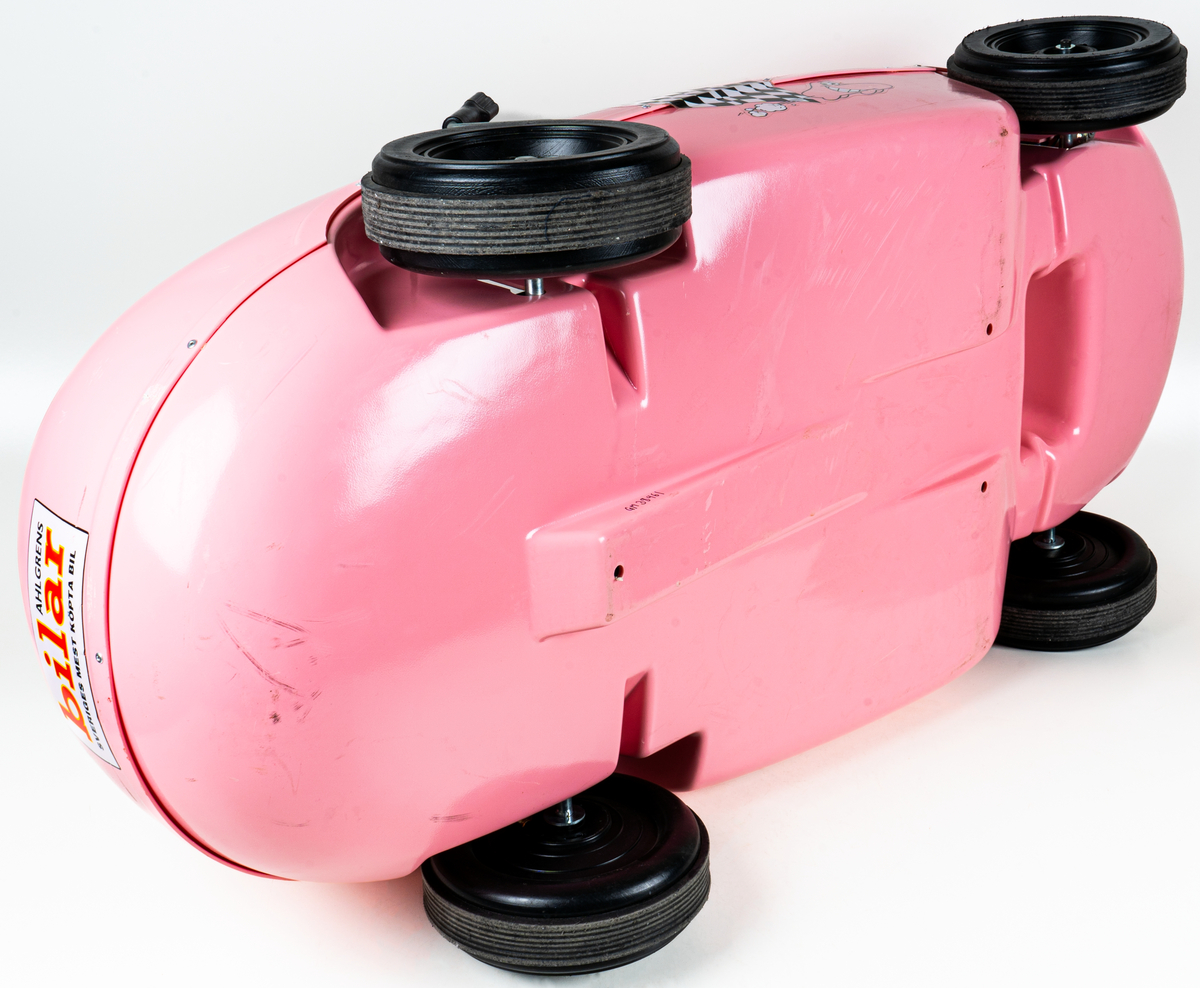 Trampbil av rosa plast, i form av en Ahlgrens bil. Fyra svarta hjul, svart ratt, dekaler på utsidan.