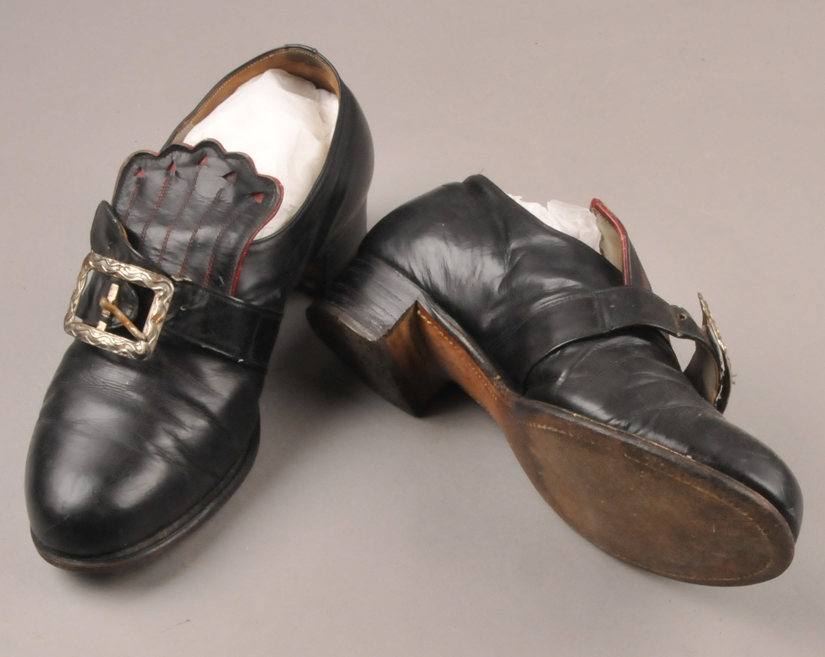 Svarte bunadsko med raud dekor på pløsa. Støpt metallspenne. Skoene har fått nye hælar, som er plugga med metallnaglar.