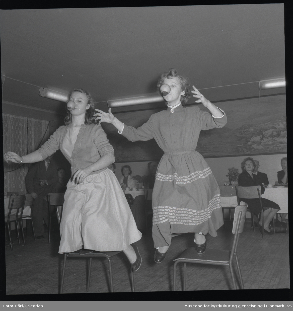 To unge damer har det moro på juletrefesten i den katolske menigheten i Hammerfest i 1957. De holder hver sin skje i munnen og balanserer epler på dem mens de går kapp over stoler. Dama til høyre er Lillian Trondsen. I bakgrunnen ser man flere personer som sitter ved bordene og ser på.