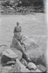 Kvinne i rutet kjole sitter på en stor stein ved bredden av 