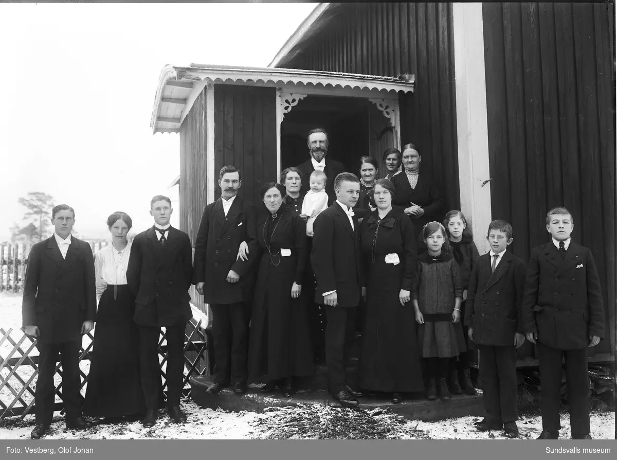 Gruppbild med vuxna och barn vid ingången till en byggnad. Möjligen en begravning, de flesta har mörka kläder, männen har vita flugor och två av kvinnorna har näsdukar i kjolslinningen.