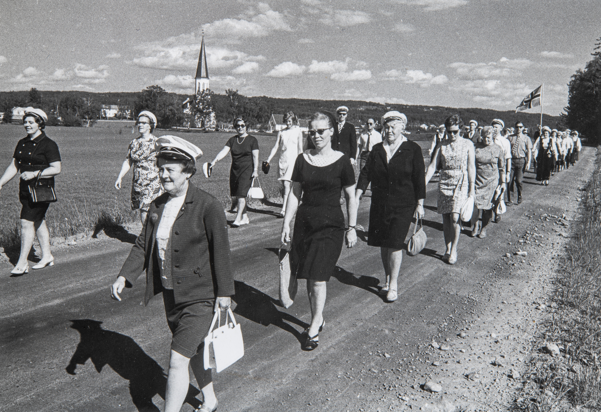 Ottestad Blandede kor masjerer i forbindelse med et sangerstevne i Romedal på 1960-tallet. Romedal kirke i bakgrunn. Nærmest kamera, i første rekke går Agnes Lund.