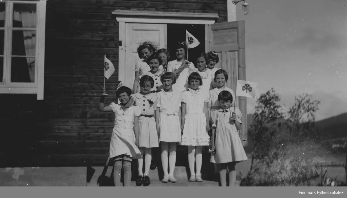 Et gruppebilde av jenter på en trapp ved døra. Noen jenter holder i hånd et flagg med NKS (Norske Kvinners Sanitetsforening). Stedet er Talvik 1939.