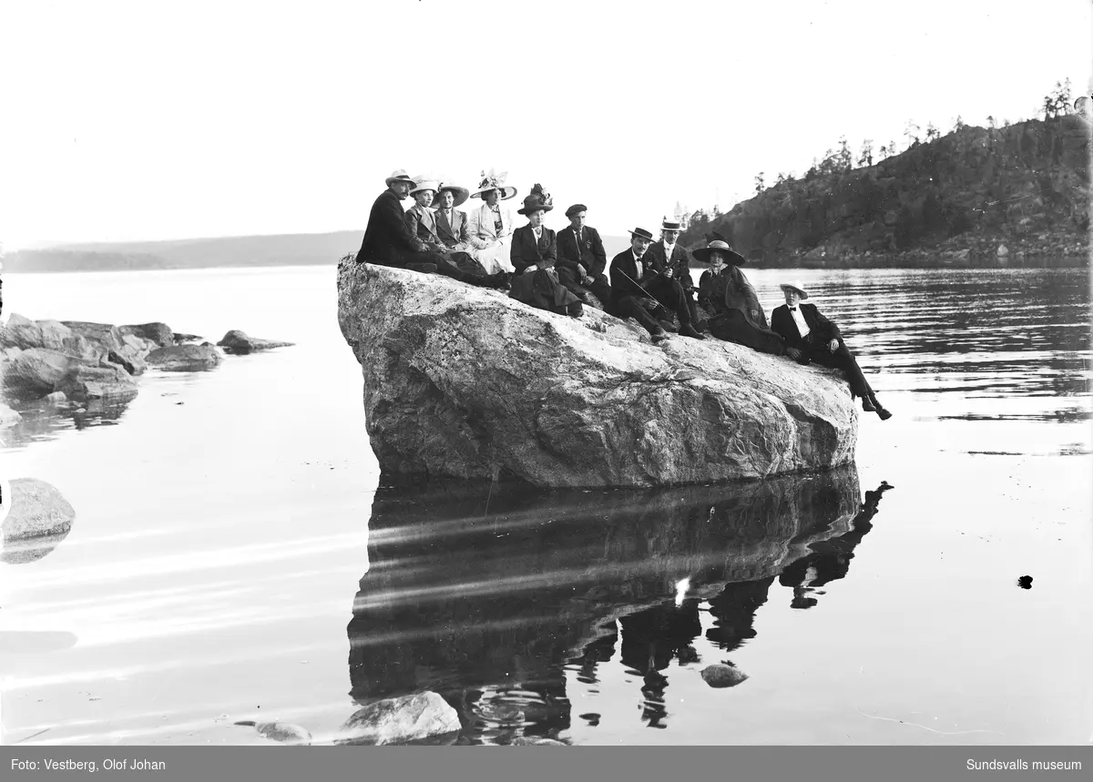 Båtutflykt till ön Skigan i Juniskärs skärgård. Tio personer sitter på en stor klippa i strandkanten, en av männen håller i en fiol.