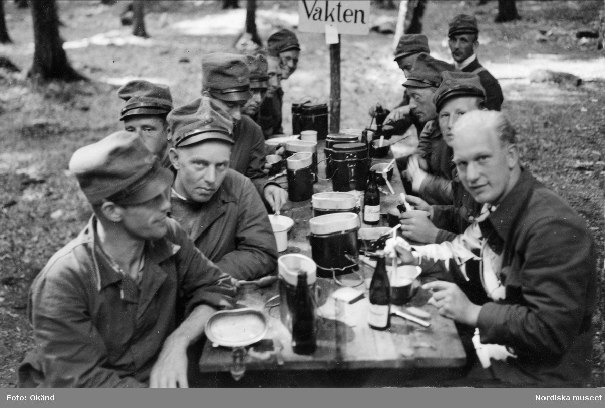 Beredskapsmän i uniform sitter utomhus vid ett dukat bord och äter en måltid.