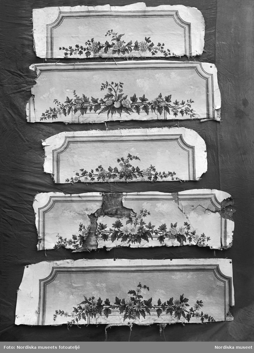 Dörröverstycken som ingår i en svit väggfält och delar av väggfält, 14 stycken, målade med limfärg på grovt papper och uppklistrade på grov, tät linneväv. Målade i en något naivistisk stil med fina detaljer. Samtliga delar med målad, profilerad ram i bronsimiterande ockratoner Från  Freluga i Hälsingland. Målningarna är utförda av Jonas Hartman ca. 1775-1804. Dörröverstycken från 1799.