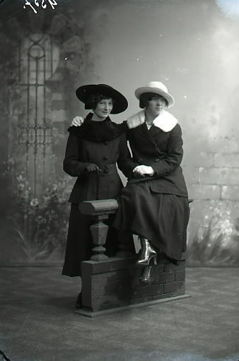 Ateljébild.  Två unga kvinnor i helfigur där en sitter på en rekvisitabalustrad. Båda iförda ytterkläder med pälskrage samt hatt. Hon till höger bär snörkängor med hög klack. Beställare: Nanny Herming, Reinholdssons, Falkenberg.