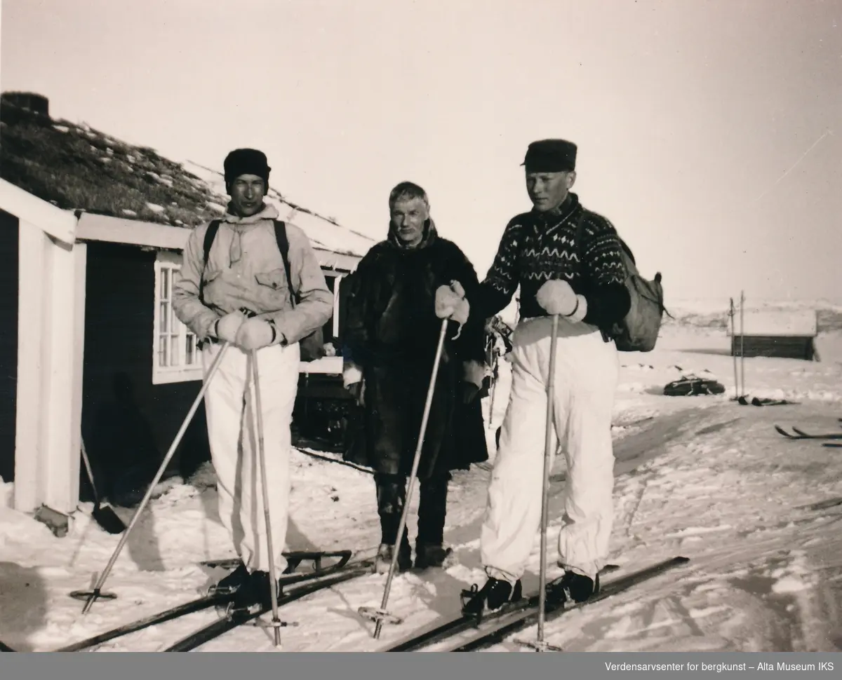 Tre menn foran ei hytte ved Joatka fjellstue. To menn har på skiutstyr, den tredje er kledd i pesk, bellinger og skaller.