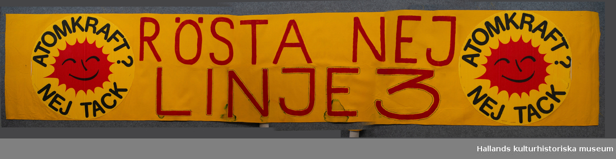 Banderoll av gult bomullstyg med antikärnkraftssymboler, (den glada solen) i wellpapp. Röd text: "RÖSTA NEJ LINJE 3".