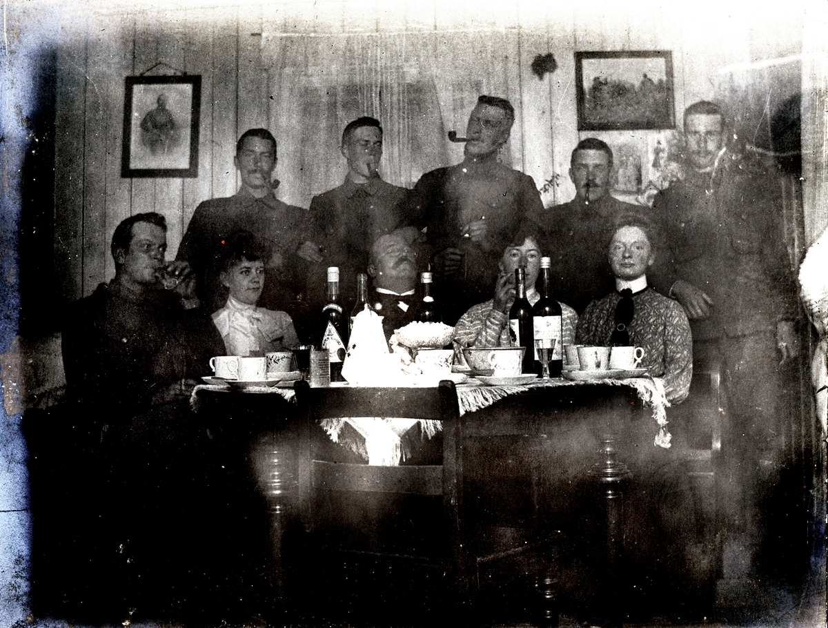 Soldater og kvinner sitter og står ved et bord med flasker og kopperr på.
Fest på Askhuset 1905.
Bilde er fra fotoalbum GM.036887.