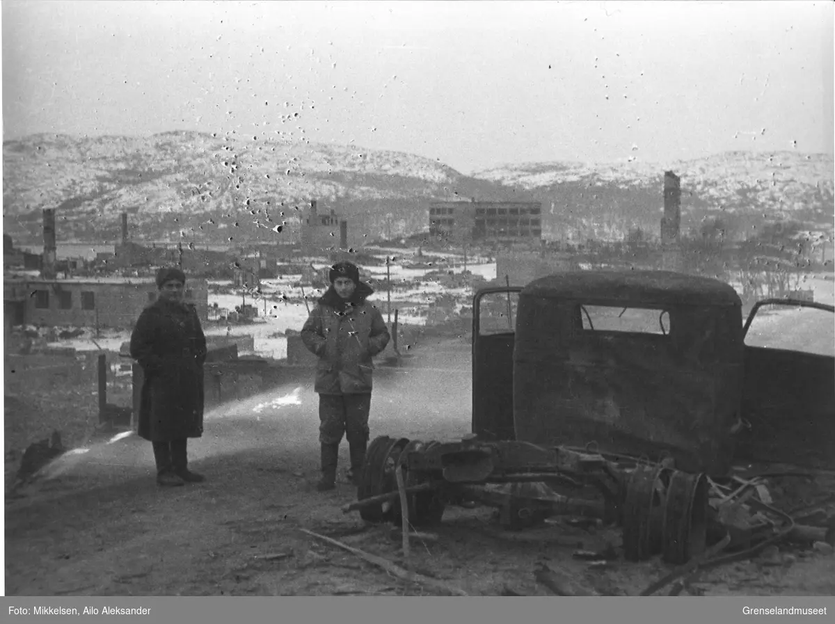 To sovjetiske soldater poserer ved en utbrent lastebil med ruinene av Kirkenes by i bakgrunnen. Bildet er tatt på høsten-vinteren i 1944 etter frigjøringen.