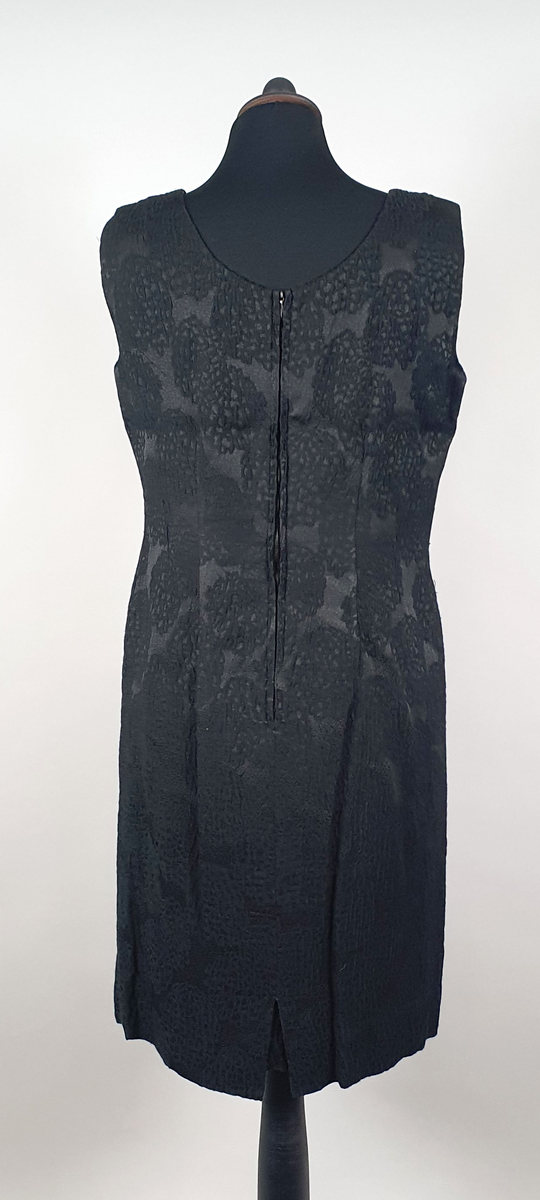 Svart kjole av syntetiske fibre, med innvevd mønster. Svarte bånd over brystet. Glidelås i ryggen. Fôr av svart kunstsilke.