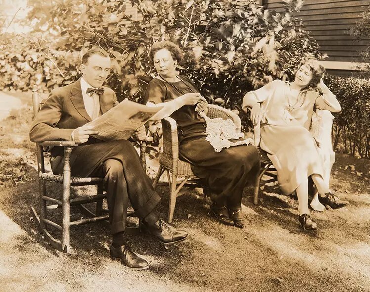 To kvinner, trolig mor og datter, og en mann i hagen. Mannen leser aviser, den eldste av kvinnene hekler. Mannen og den eldre kvinnen har runde briller. Bildet er trolig fra 1920- årene.