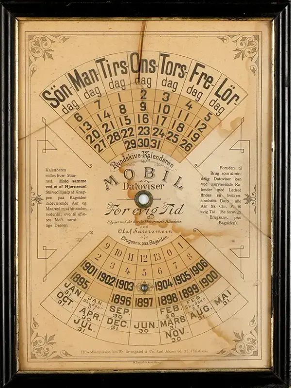 En avlang evighetskalender i papp i en mørk treramme. Kalenderen har en rund skive som kan dreies for å vise korrekt dato. Fint dekorert med grafiske elementer fra tidlig 1900-tall.