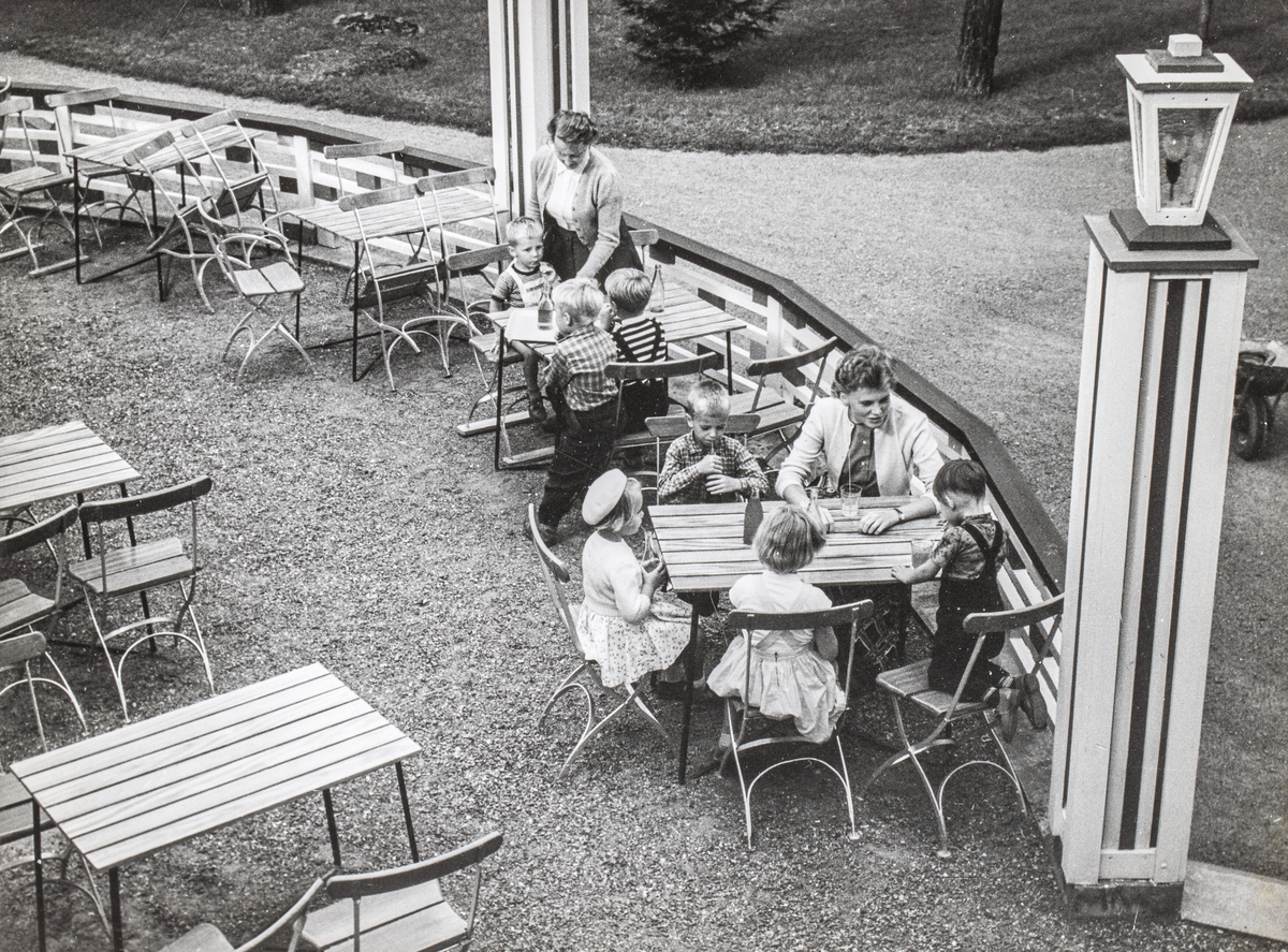 Uteservering i Folkets Park i Linköping. 1950-60-tal. Vid två av borden sitter barn, vid det ena står en kvinna och vid det andra sitter en kvinna.