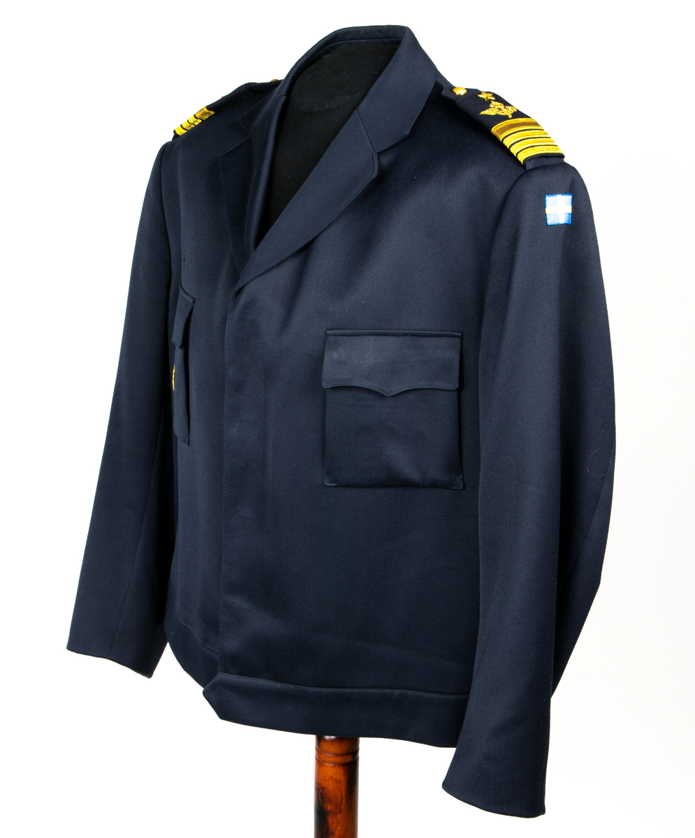 Jacka m/87. Blå uniformsjacka med dold knäppning med fyra knappar av plast. Jackan har midjelinning, fasta axelklaffar vilka försetts med försetts med gradbeteckning, överste 1 graden.  På vänster överarm är en nationsbeteckning i form av en svensk flagga fastsydd och på höger bröstficka sitter försvarsstabsmärke m/49.