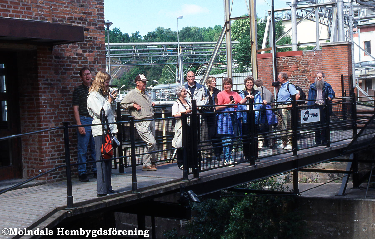 Amerikaner på besök i Mölndals Kvarnby, år 2002. De står på gångbron över forsen vid industribyggnaden Stora Götafors. Lars Gahrn gör dem sällskap.