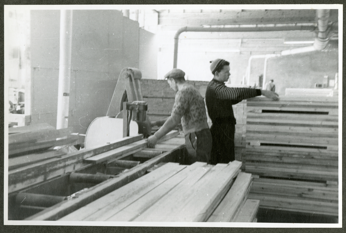 Foto av paller med tremateriale og arbeidere i verkstedmiljø ved Aall-Ulefoss Brugs Dørfabrikk