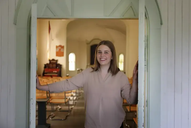 I sommer kan du få guidet tur i friluftsmuseet. På bildet kan man se en ung kvinne som smiler og står i døråpningen på et antikvarisk hus ved Norsk utvandrermuseum.