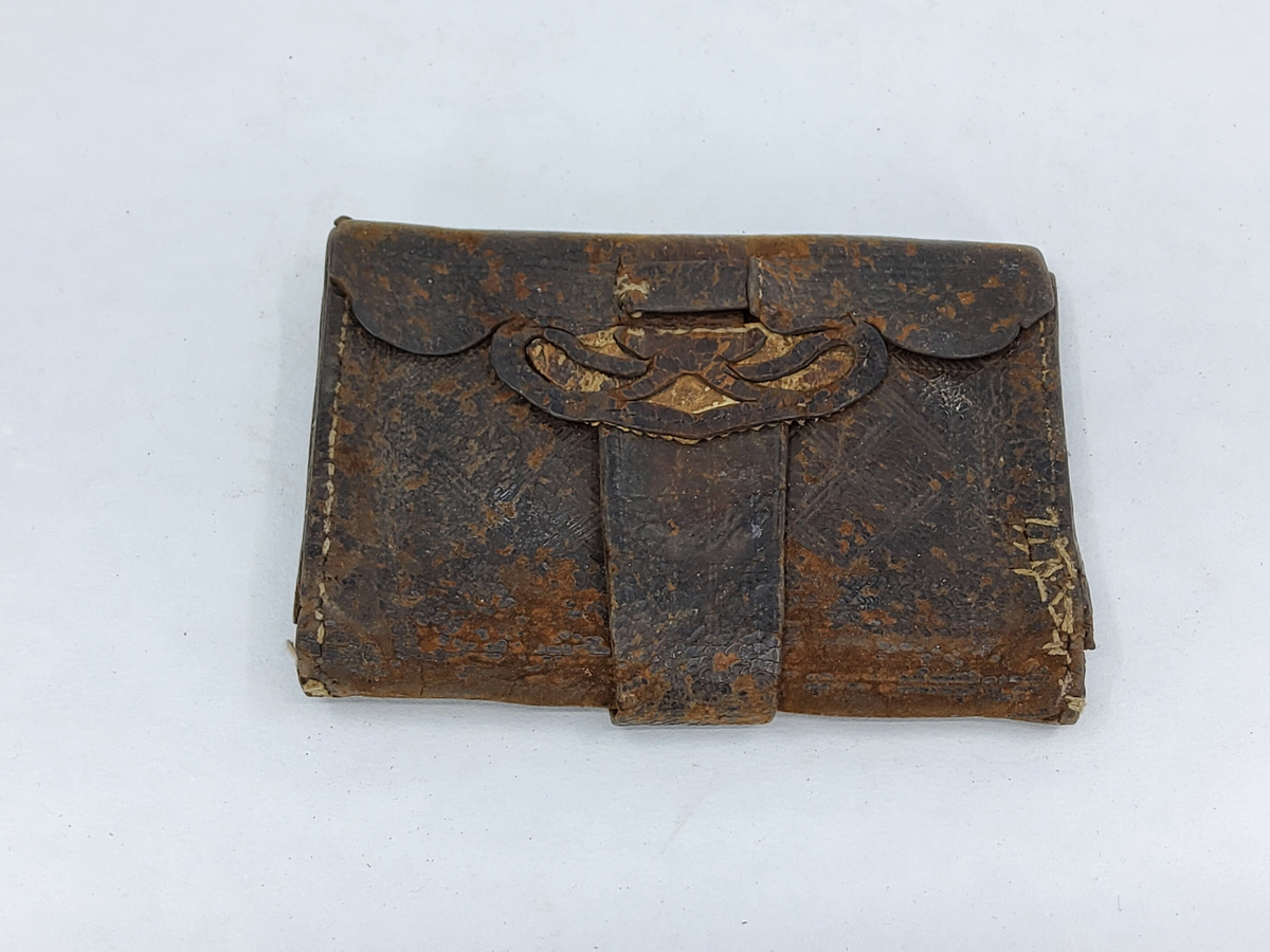Plånbok av brunt skinn med pressade mönster, dubbel, med många olika fack, utskärningar på framsidan, lång rem för låsning. Sliten.