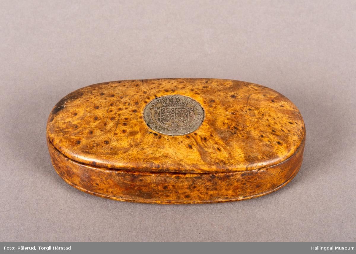 En brun, oval snusdåse i valbjørk. Lokket er festet med trehengsler på den ene langsiden. Innfelt i lokket sitter en 12-skilling fra 1719. Under bunnen er det innskåret "K O S ANO 1642".
