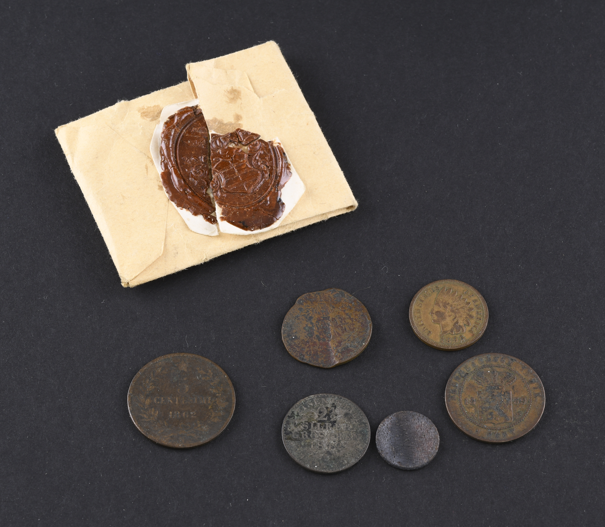 Seks mynter i varierende størrelse og tilstand. Myntene er fra USA, Preussen, Italia og Nederland. Myntene lå oppbevart i en papirpose med rød vokssegl.