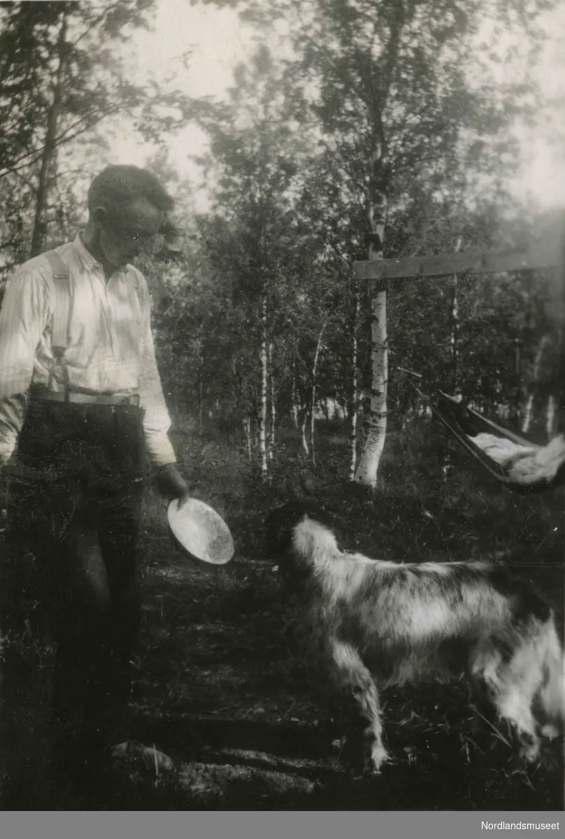 En mann holder en skål mot en hund. I bakgrunnen ser vi en hengekøye.
