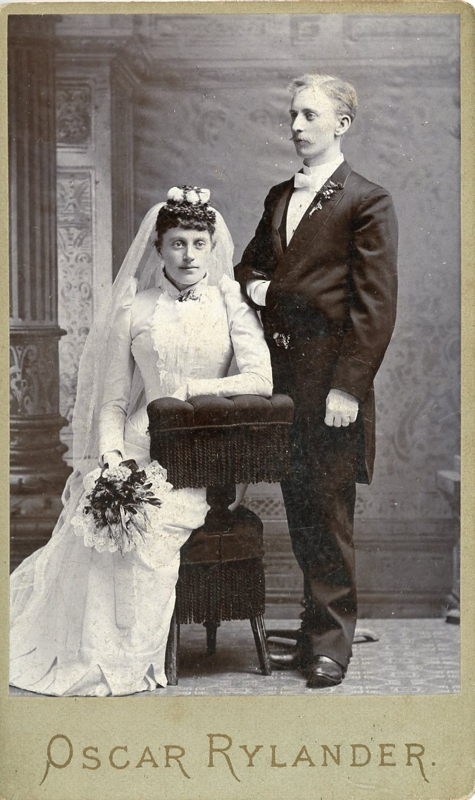 Ett brudpar. Kvinnan sitter på en pall. Mannen står i Napoleon-pose.
