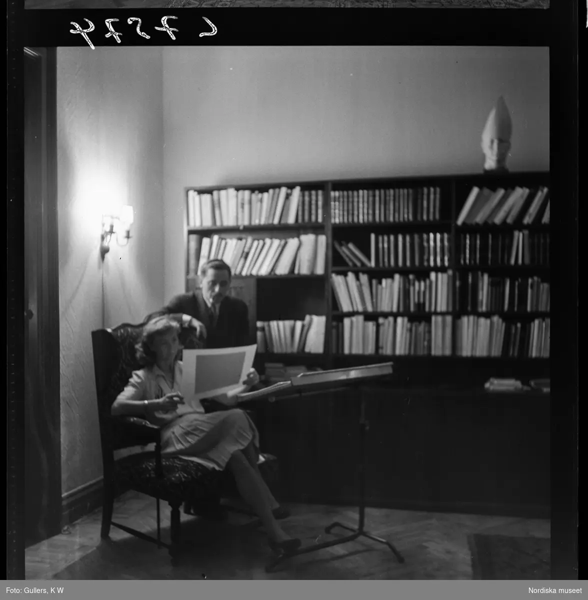 513 D Spooners Hemma hos redaktör Karlströmer. Man och kvinna tittar på en bild sittandes framför en bokhylla i ett vardagsrum.